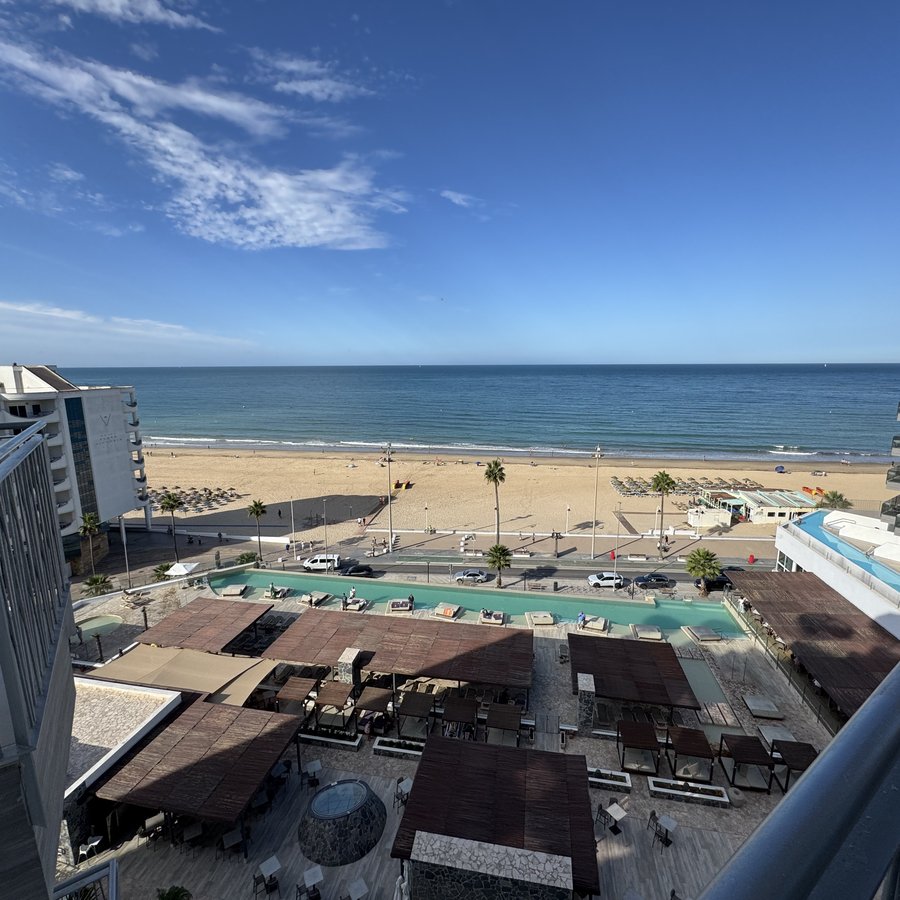 Blick aus dem achten Stock über Hoteldächer und Poolterrassen hinweg auf den Strand und das Meer, die Horizontlinie scharf abgesetzt vor klarem Himmel.