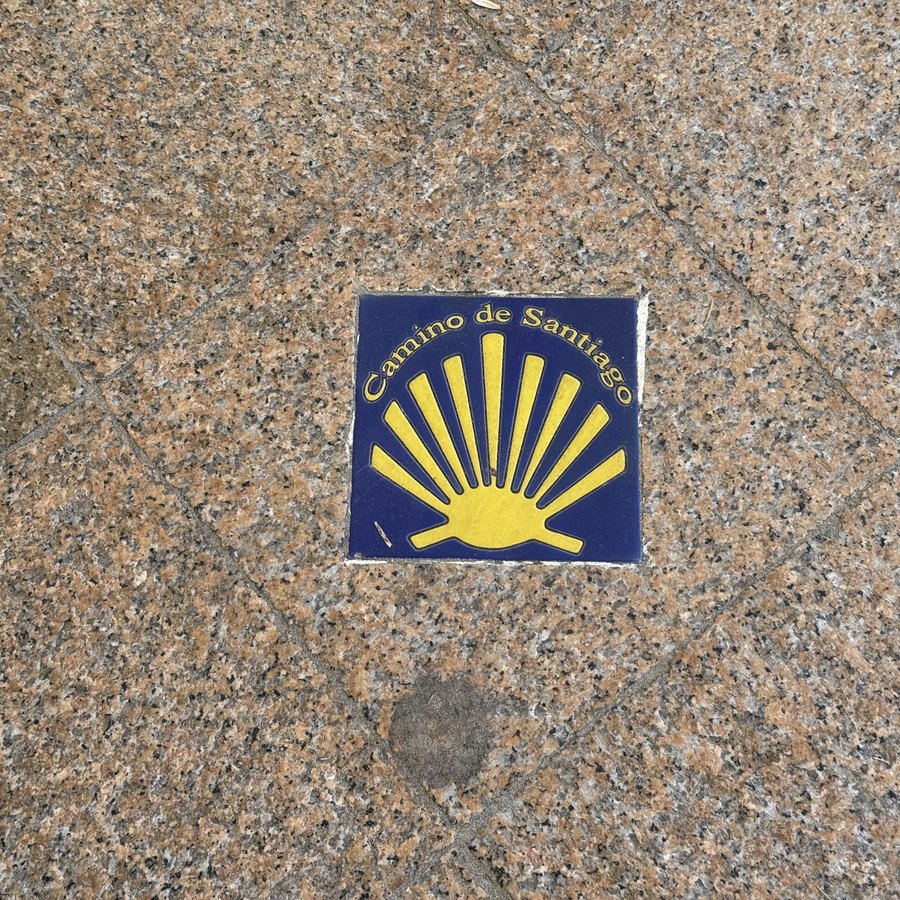 Eine blaue Bodenkachel im Gehweg mit der gelben Jakobsmuschel und der Aufschrift Camino de Santiago.