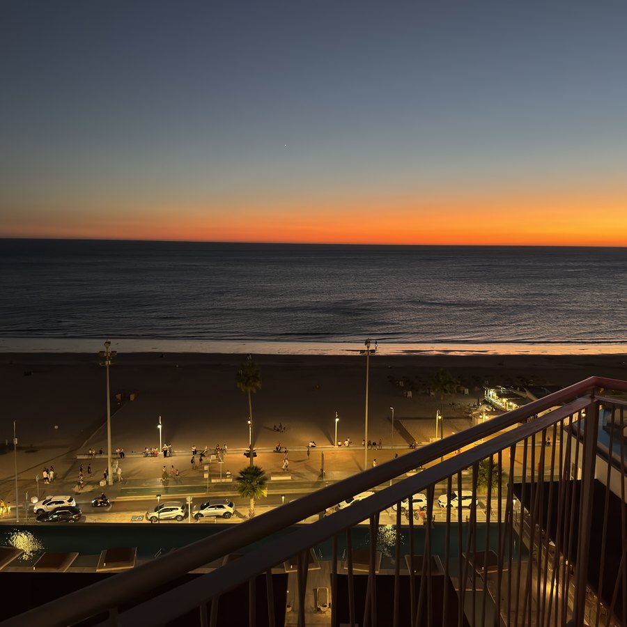 Sonnenuntergang über dem Atlantik vom Balkon aus: oranger Himmelsstreifen über dunklem Meer, darunter die beleuchtete Strandpromenade.
