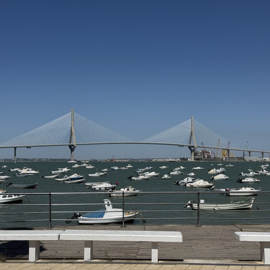 Blick über die Bucht von Cádiz auf die weiße Schrägseilbrücke, davor ankern zahlreiche kleine weiße Boote im flachen Wasser.