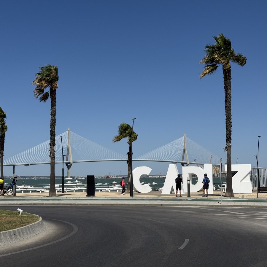 Große weiße Buchstaben bilden den Schriftzug CÁDIZ an einer Straße, dahinter Palmen und die Schrägseilbrücke.