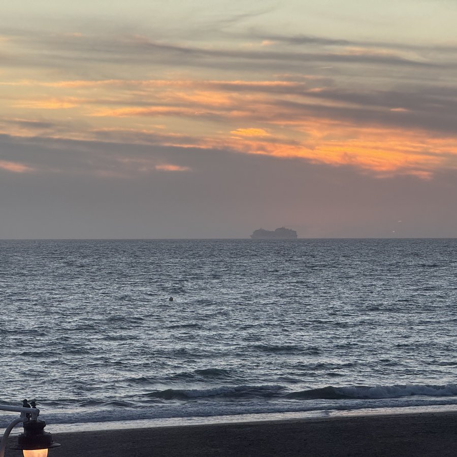 Sonnenuntergang über dem Meer in Rosa und Grau, am Horizont die kleine Silhouette eines großen Kreuzfahrtschiffs, im Vordergrund der dunkle Strand.