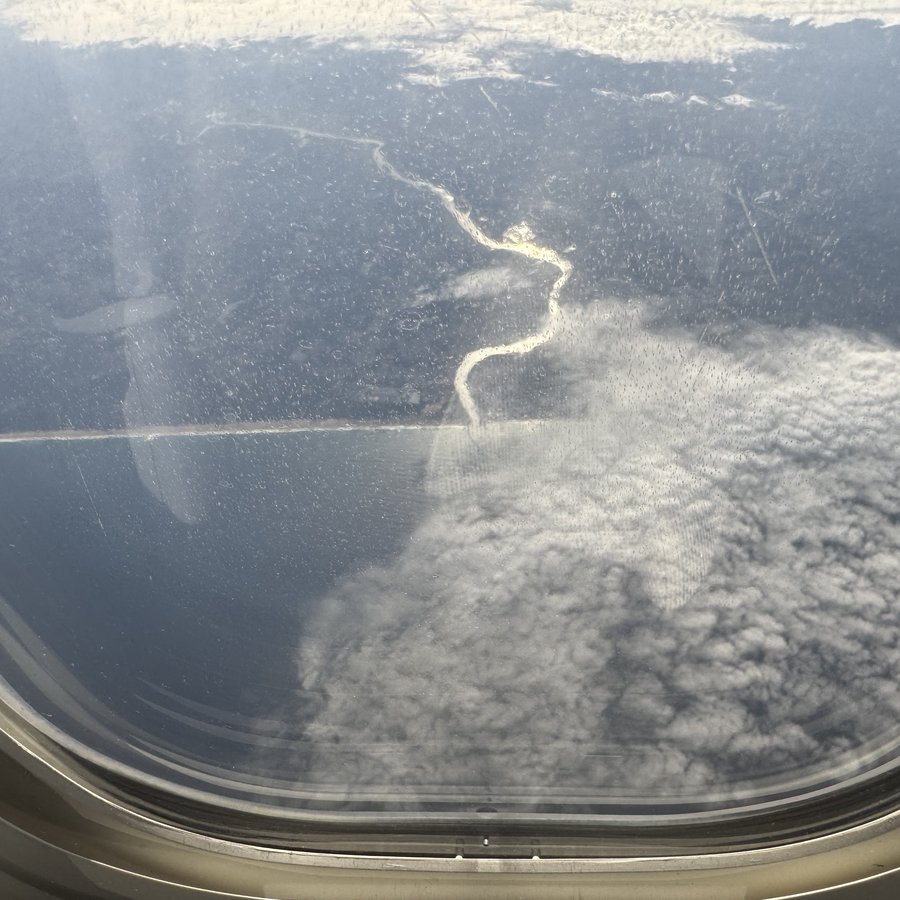 Blick durch das ovale Flugzeugfenster auf eine Küstenlinie weit unten: hellbrauner Sand, davor das Meer, darüber eine geschlossene Wolkendecke.