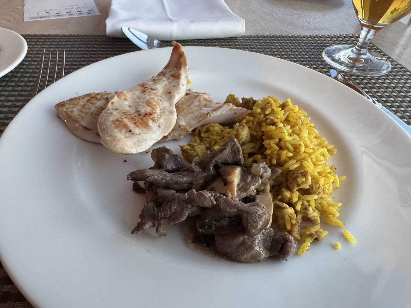 Ein Teller mit einem Stück gegrilltem Hähnchen, dunklem Fleisch in Sauce und gelbem Safranreis, daneben ein Weinglas.
