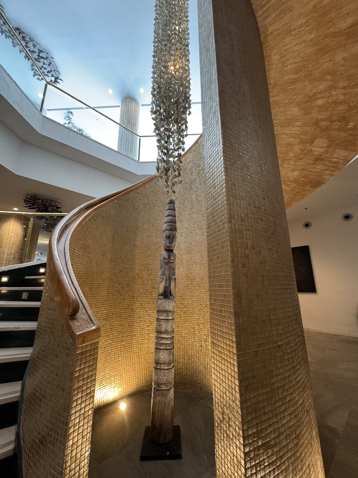 Im hohen Atrium des Hotels hängt ein schmaler, mehrere Stockwerke langer Kristallleuchter neben einer geschwungenen Treppe an einer goldfarbenen Wand.
