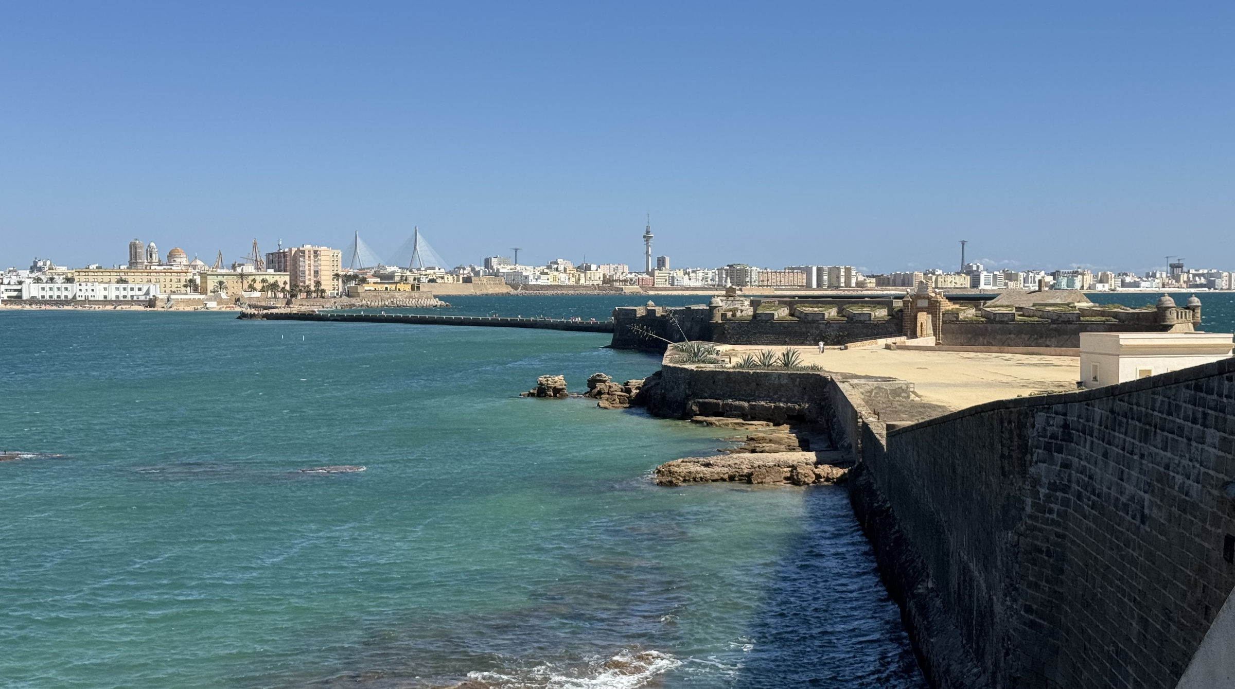 Blick über türkisfarbenes Wasser auf die Silhouette von Cádiz: eine flache Stadtzeile am Horizont, davor eine Landzunge mit heller Ufermauer und dunklen Felsen im Wasser.